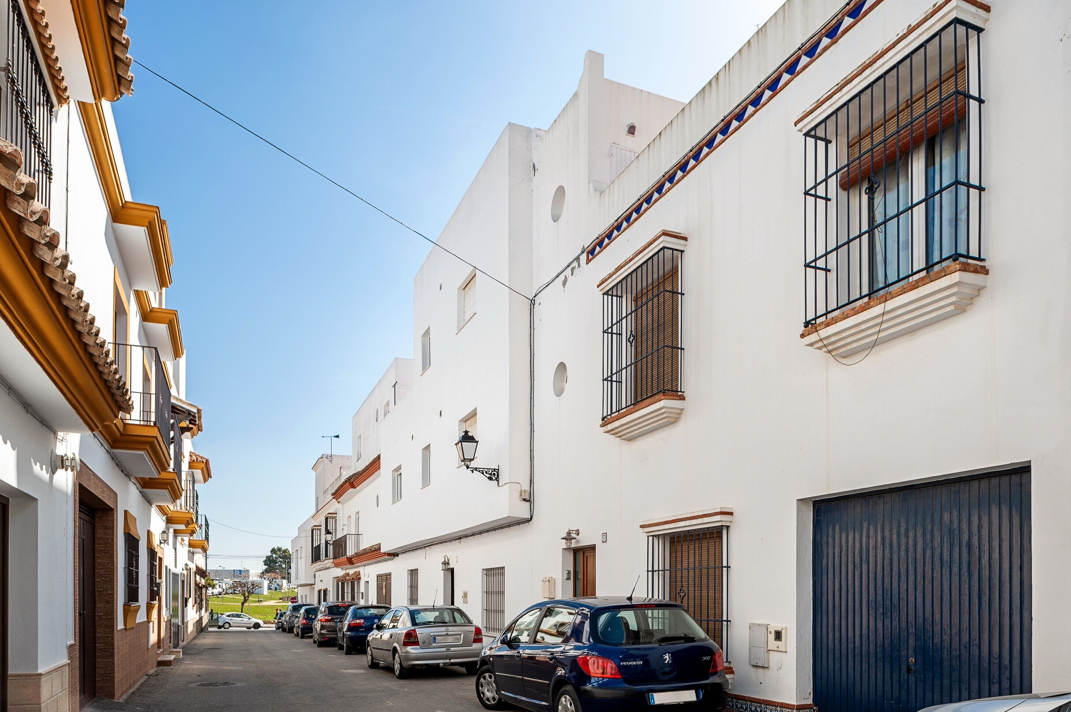 Una calle estrecha bordeada de edificios blancos, coches aparcados y un cielo azul despejado.