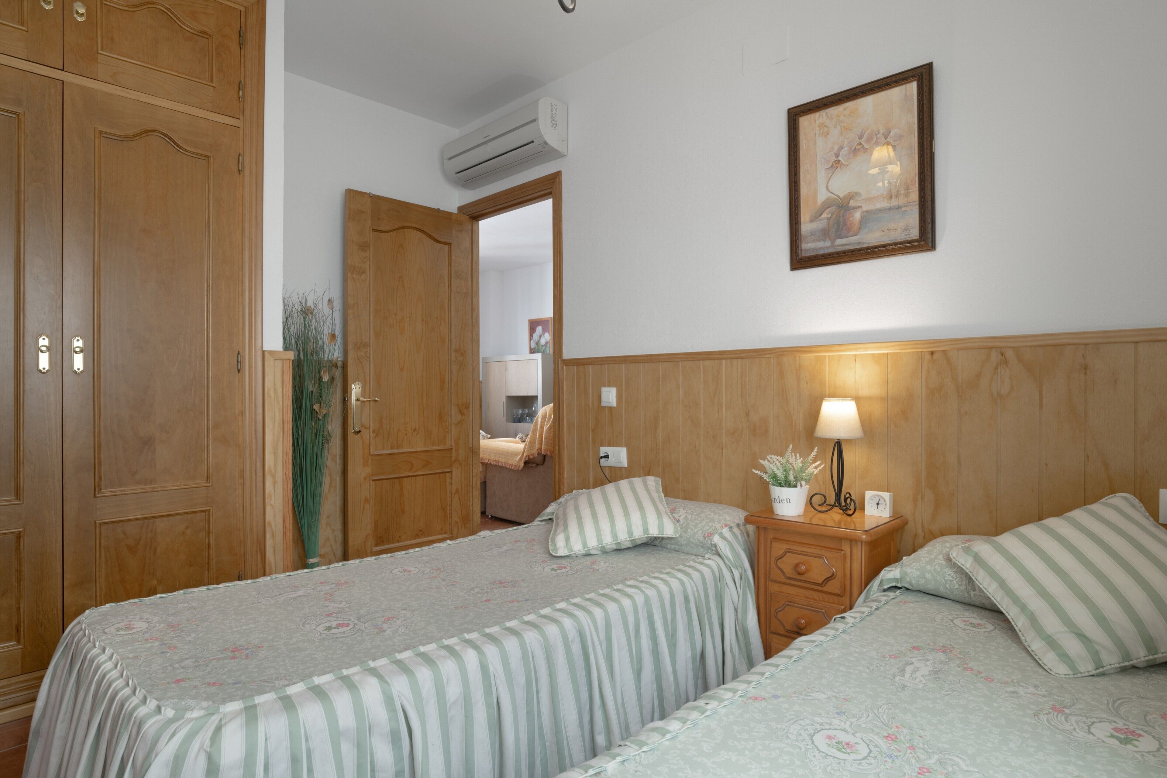 Slaapkamer met twee bedden, een houten kledingkast, een nachtkastje met een lamp en een wandairconditioner.