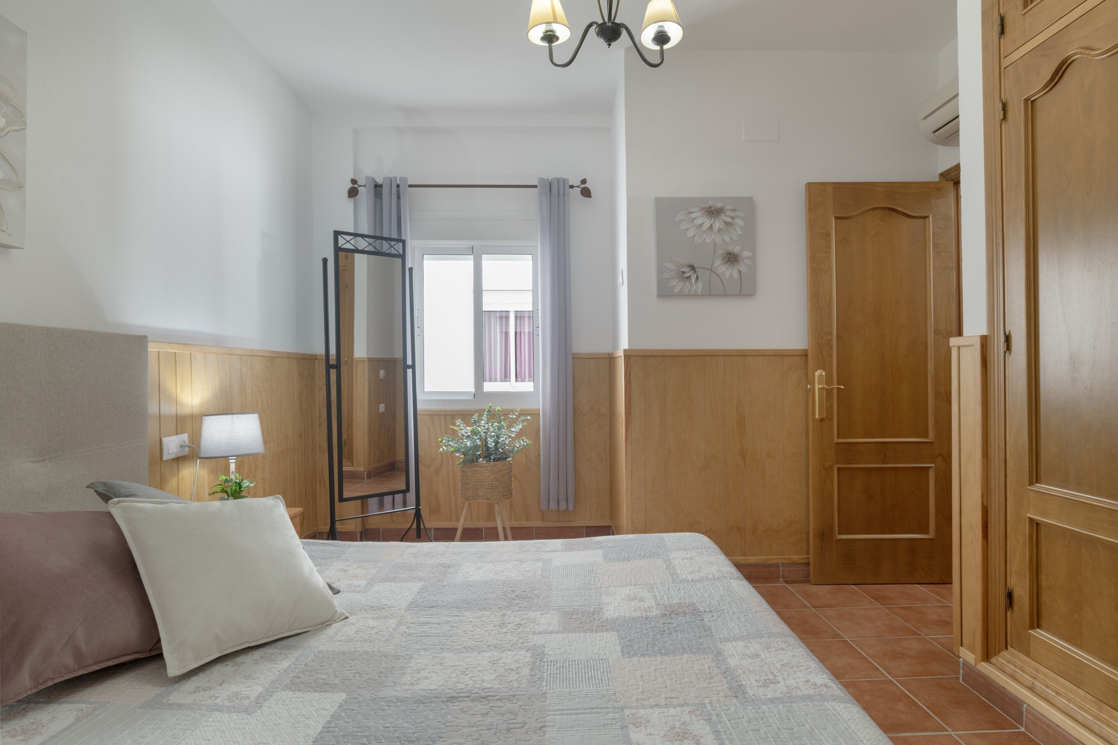 A bedroom with a bed, a wooden wardrobe, a mirror, a window with curtains, and a wall-mounted lamp.
