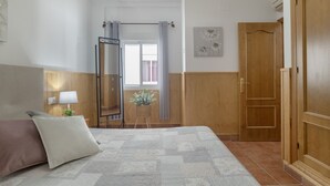 A bedroom with a bed, a wooden wardrobe, a mirror, a window with curtains, and a wall-mounted lamp. - Family Friendly Apartment "Rocio" with Terrace, Air Conditioning and Wi-Fi (Conil de la frontera)
