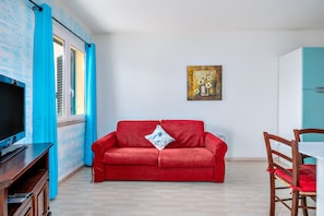Un sofá rojo, un televisor, un cuadro, una silla de madera y un mueble azul verdoso en una habitación con suelo de baldosas.