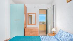 Un dormitorio con cama, armario, cómoda, espejo, mesita de noche con lámpara y puerta corrediza al balcón.