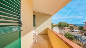Un balcón con un persiano verde, una silla blanca y vistas a una calle y edificios.