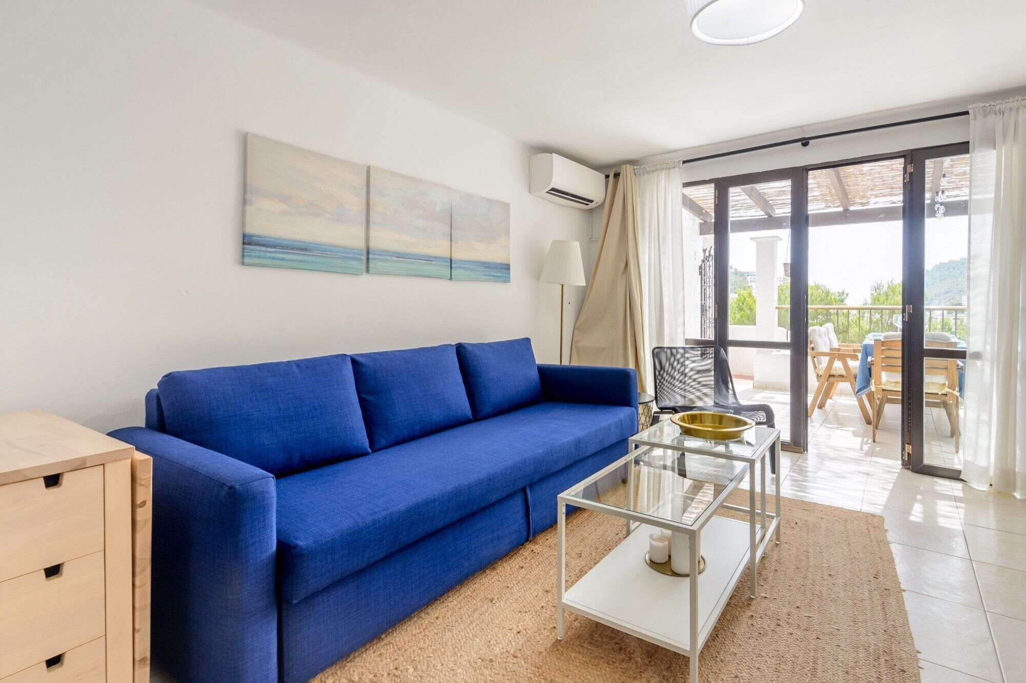 A living room with a blue sofa, a glass coffee table, a lamp, and a view of the outdoors.