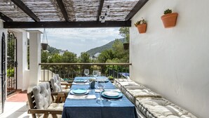 A dining area with a table set for four, a cushioned bench, and a view of greenery and hills. - Holiday Home "Casita en Cala Llonga" with Sea View, Shared Garden & Pool (Cala Llonga)