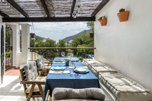 Un coin repas avec une table dressée pour quatre personnes, un banc rembourré et une vue sur la verdure et les collines.
