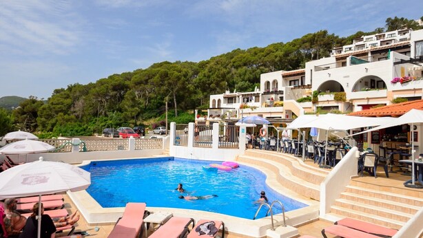 A swimming pool with lounge chairs and umbrellas, surrounded by buildings with balconies and a forested hillside. - Holiday Home "Casita en Cala Llonga" with Sea View, Shared Garden & Pool (Cala Llonga)
