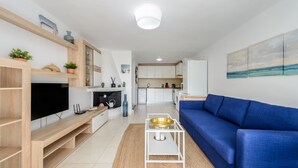 A modern living room with a blue sofa, a flat-screen TV, wooden shelves, and a kitchen in the background. - Holiday Home "Casita en Cala Llonga" with Sea View, Shared Garden & Pool (Cala Llonga)