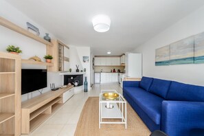A modern living room with a blue sofa, a flat-screen TV, wooden shelves, and a kitchen in the background. - Holiday Home "Casita en Cala Llonga" with Sea View, Shared Garden & Pool (Cala Llonga)