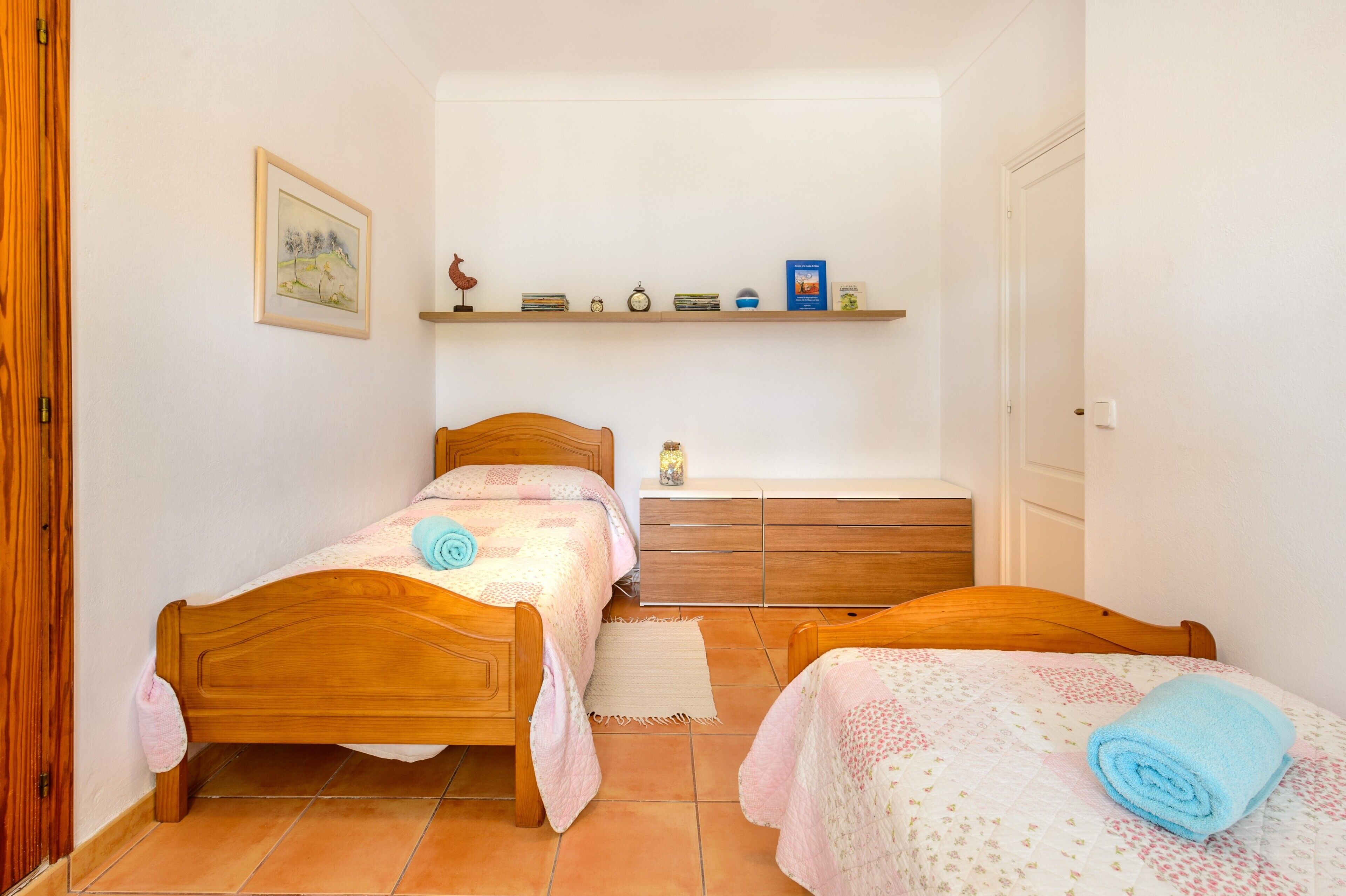Un dormitorio con dos camas de madera, una mesita de noche, un aparador y una estantería con libros y objetos decorativos.