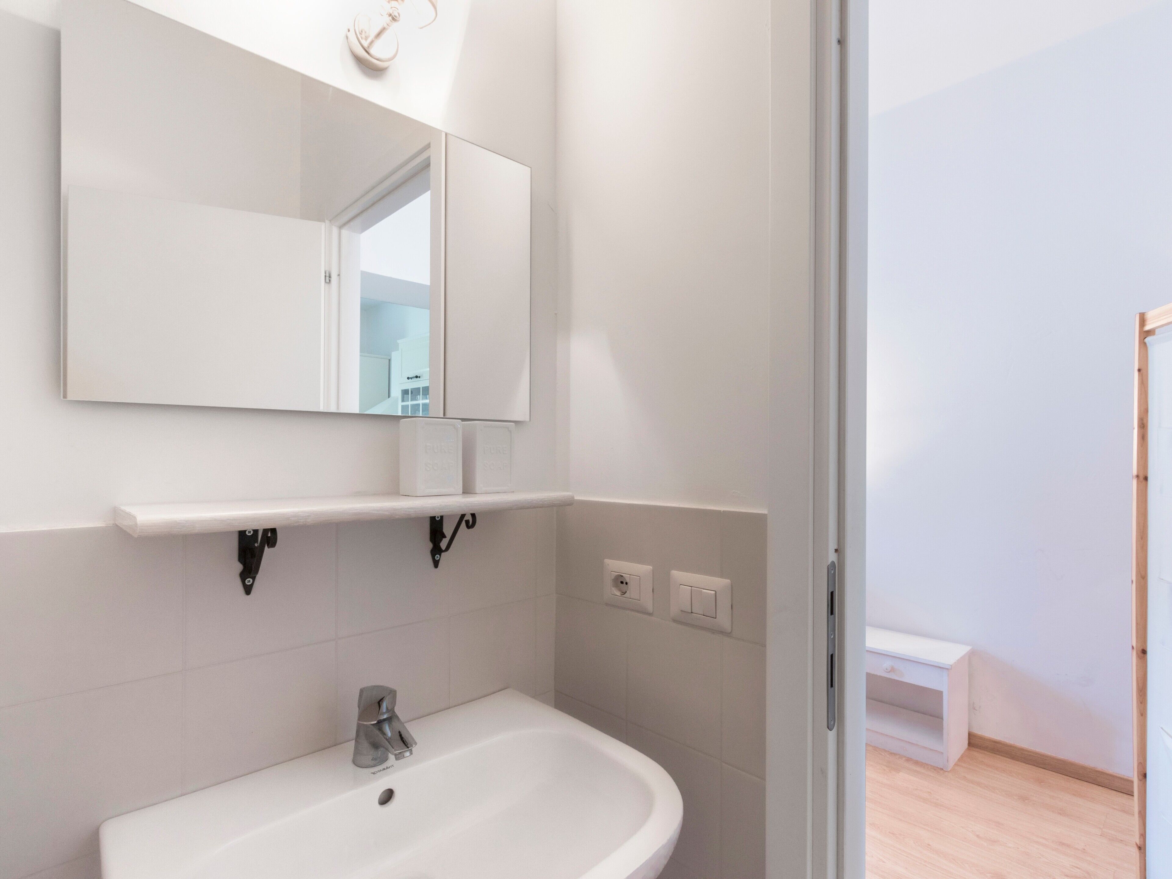 Une salle de bain avec un lavabo blanc, un miroir et une étagère au-dessus du lavabo.