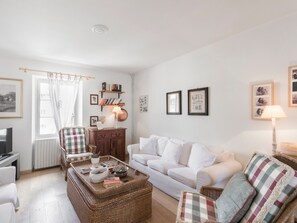 A living room with a white sofa, a wicker coffee table, a bookshelf, and a window with curtains.