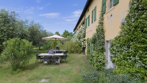 A patio with a table and chairs, surrounded by greenery and a building with green shutters.
