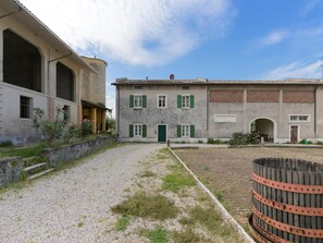 A courtyard with a building featuring a green door and shuttered windows, a stone wall, and a gravel path.