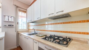 Une cuisine avec des meubles blancs, une cuisinière à gaz, un évier et une fenêtre avec des stores.
