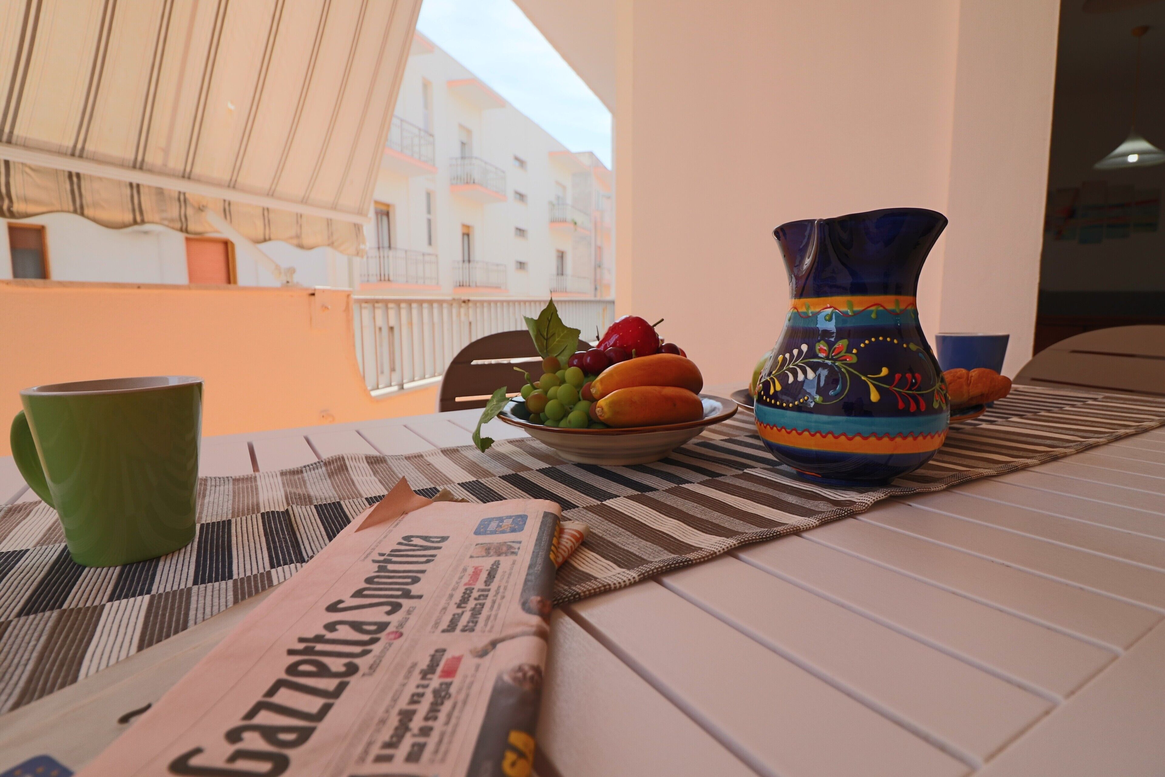 A table with a newspaper, a bowl of fruit, a vase, and a green mug.