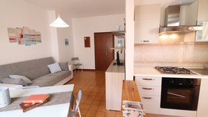 Een moderne keuken met een fornuis, oven en witte kasten. Er is een eethoek met een tafel en stoelen, en een grijze bank in de woonkamer.