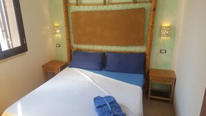Un lit recouvert d’une couverture blanche et bleue, avec deux oreillers bleus et une serviette bleue posée sur le lit. On y voit deux tables de chevet avec des lampes et une fenêtre sur le côté gauche.