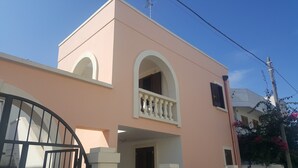 Un bâtiment rose avec une entrée fermée par un portail, un balcon orné de balustres et une petite fenêtre avec des volets.