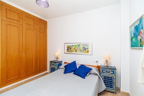 A bedroom with a large wardrobe, a bed with blue pillows, two bedside tables with lamps, and a colorful painting on the wall.