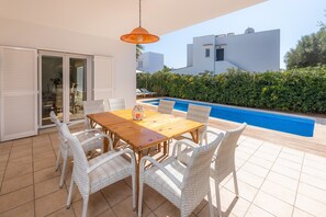 Une terrasse avec une table en bois et des chaises en osier, une piscine et une vue sur un bâtiment blanc.