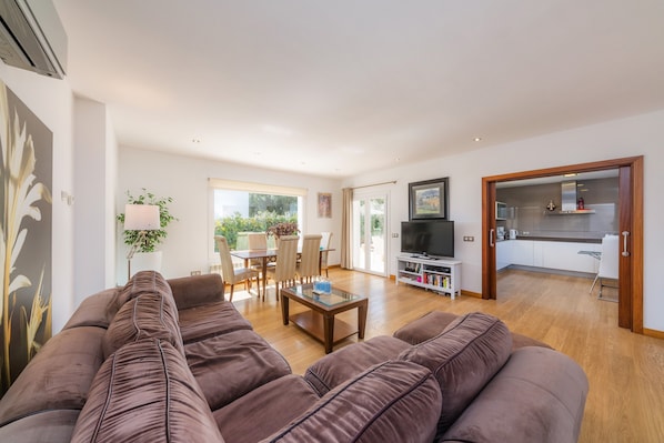 Un vaste espace de vie comprenant un canapé, une table basse et un téléviseur. On y trouve également un coin repas avec des chaises et une table, ainsi qu’une cuisine visible à travers une porte ouverte.