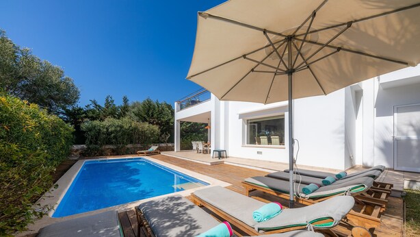 A swimming pool with lounge chairs and an umbrella, surrounded by greenery and a modern building. - House with Mountain Views, Pool, Garden, Terrace, A/C and Wi-Fi (Cala d'Or)