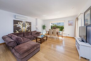 Un salon moderne comprenant un canapé d’angle, une table basse, un téléviseur posé sur un meuble et un coin repas avec des chaises et une table.