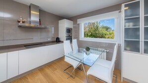 A modern kitchen with a glass dining table, white cabinets, stainless steel appliances, and a view of greenery outside. - House with Mountain Views, Pool, Garden, Terrace, A/C and Wi-Fi (Cala d'Or)