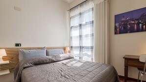 A bedroom with a bed, a desk, a chair, a window with curtains, and a wall-mounted painting.