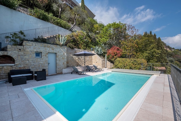 A rectangular swimming pool with a stone wall and a door, surrounded by lounge chairs and an umbrella.