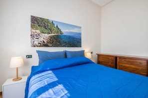A bedroom with a bed, a nightstand with a lamp, a dresser, and a wall-mounted picture of a coastal landscape.