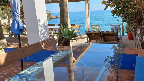 Un patio con una mesa de vidrio, sillas de madera y vista al mar.