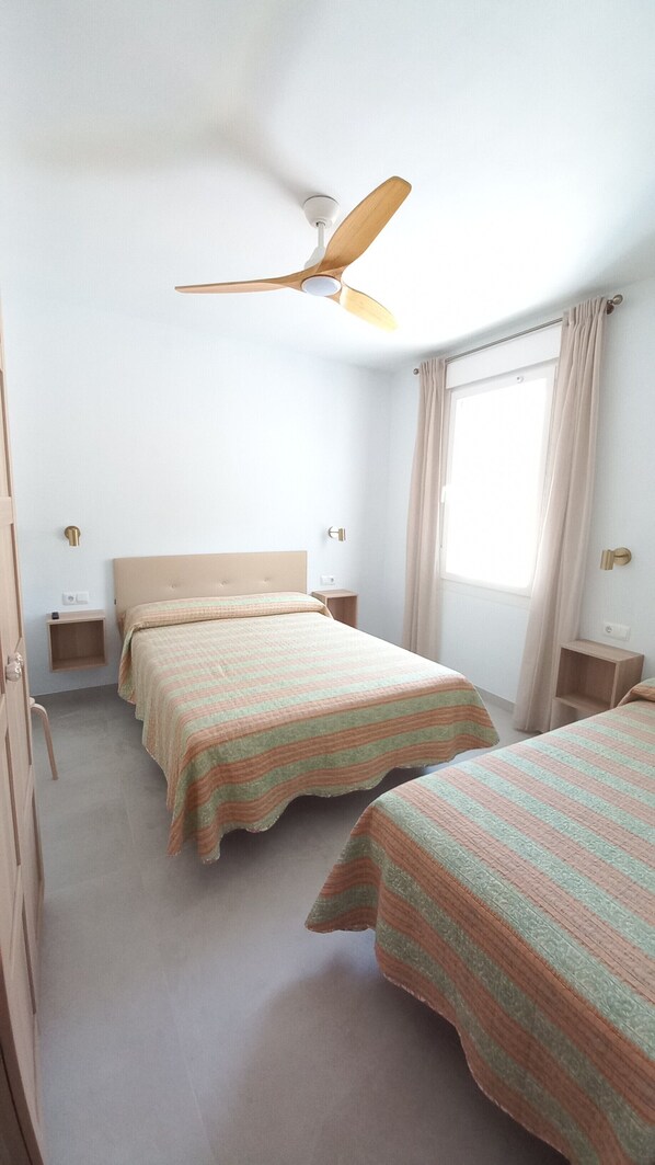 A hotel room with two beds, a ceiling fan, a window with curtains, bedside tables, and a door.