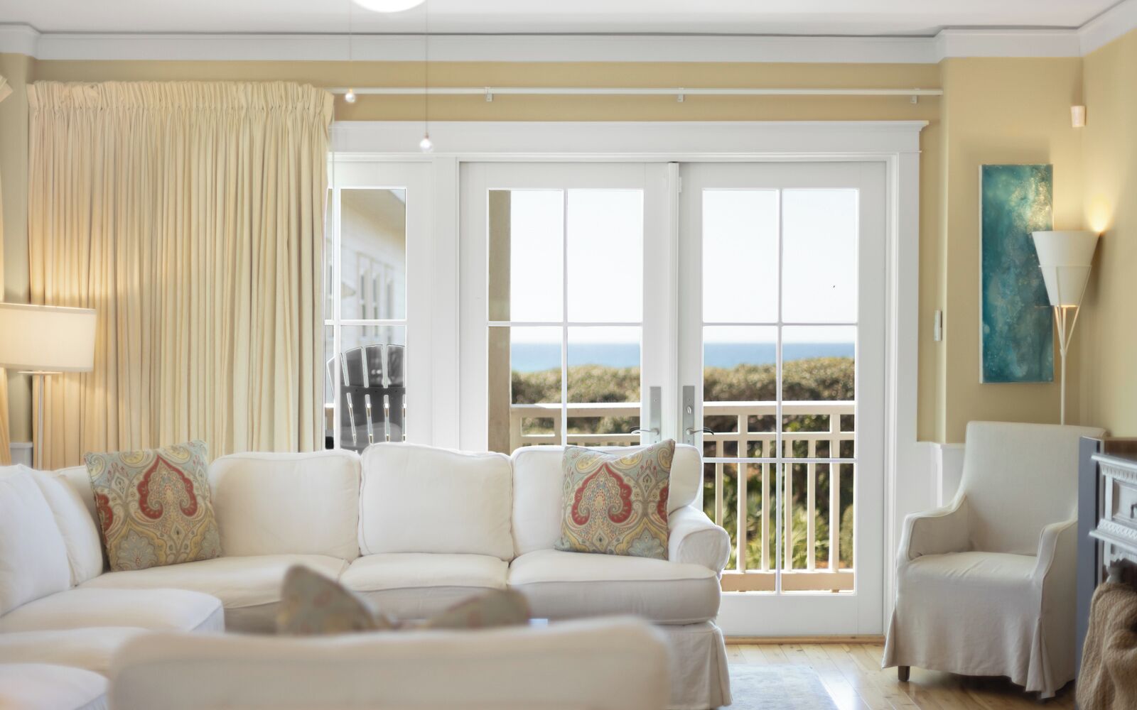 A living room with a white sofa, a chair, a balcony with a view, and a painting on the wall.