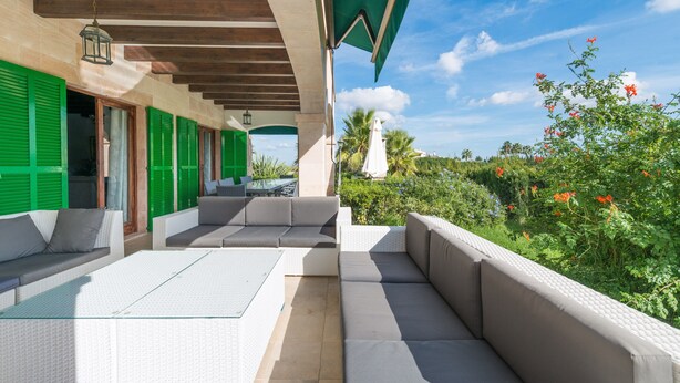 An outdoor patio with white wicker furniture, green shutters, and a view of the garden.