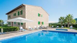 A swimming pool with lounge chairs, a stone building with green shutters, and palm trees in the background.