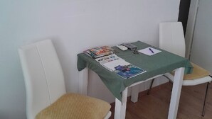 Una habitación pequeña con una televisión montada en la pared, una mesa cubierta con mantel verde y dos sillas blancas con cojines trenzados en los asientos.