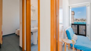 Un balcón con vistas a edificios y una piscina, una cama con cubrecama azul, un lavabo y una silla.