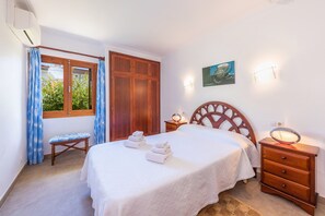 A bedroom with a bed, a wooden wardrobe, a bench, a bedside table with a mirror, and a wall-mounted light fixture.