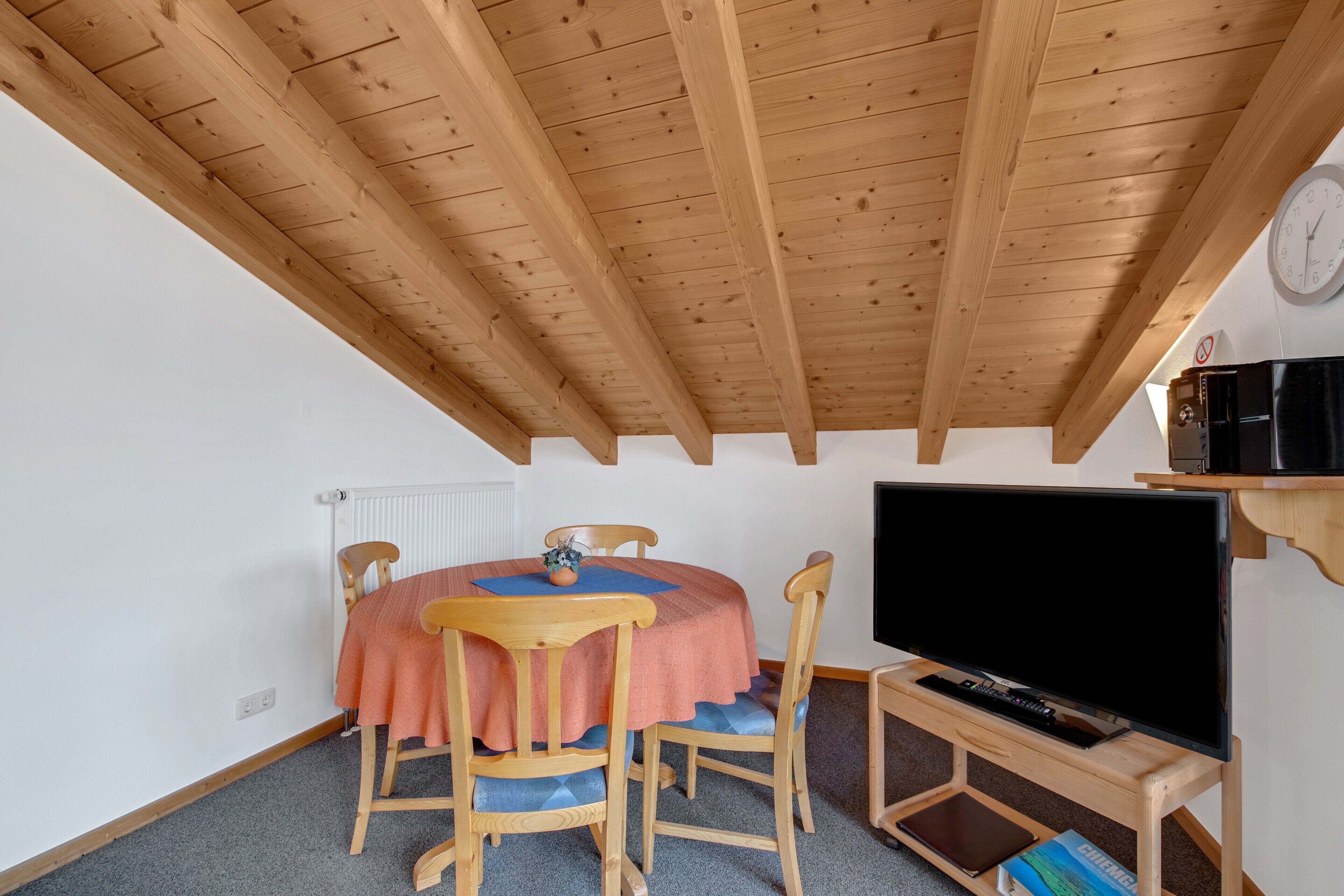 A room with a wooden ceiling, a round table with chairs, a television on a stand, and a clock on the wall.
