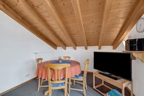 Une pièce avec un plafond en bois, une table ronde entourée de chaises, un téléviseur posé sur un meuble et une horloge au mur.