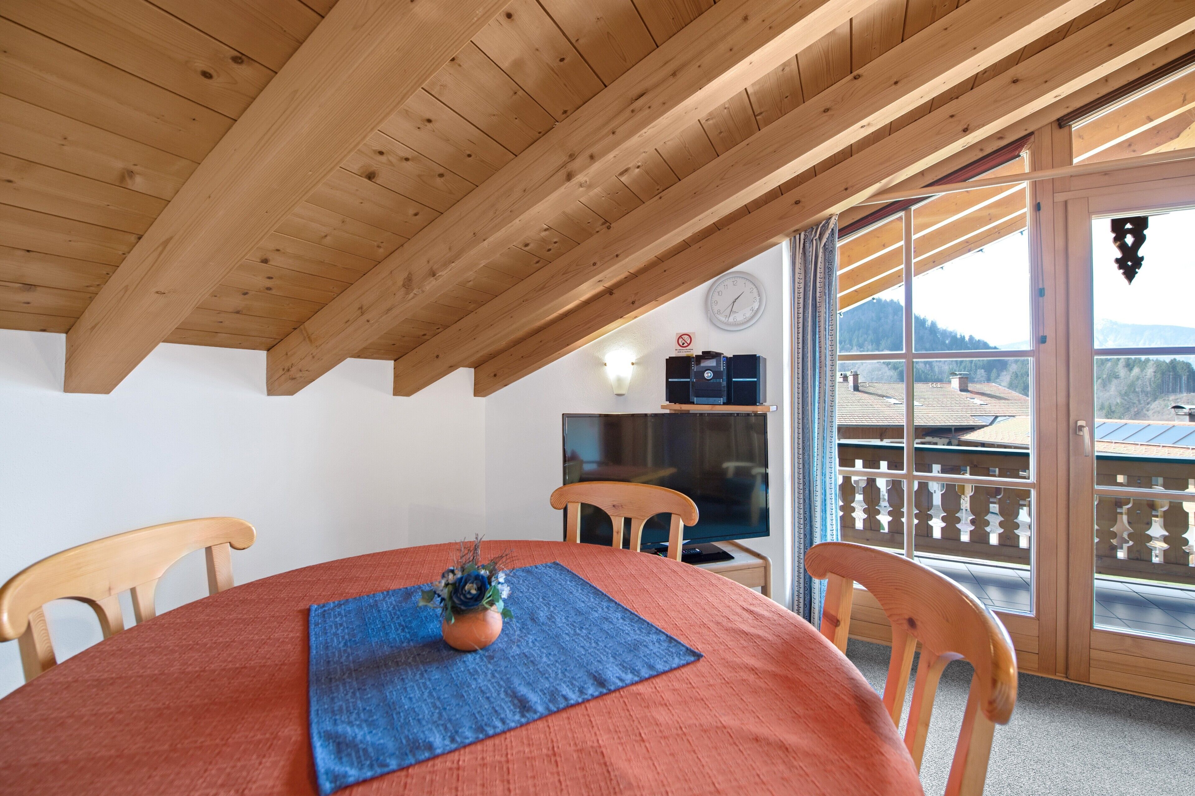 Eine Holzbalkendecke, ein Esstisch mit Stühlen, ein Fernseher und ein Balkon mit Blick.