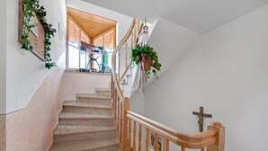 A wooden staircase with a handrail, a wall-mounted cross, and a window with a view of a room with a sewing machine. - Charming Apartment “Petrushof Rechenberg” with Mountain View, Wi-Fi, Balcony (Oberwössen)