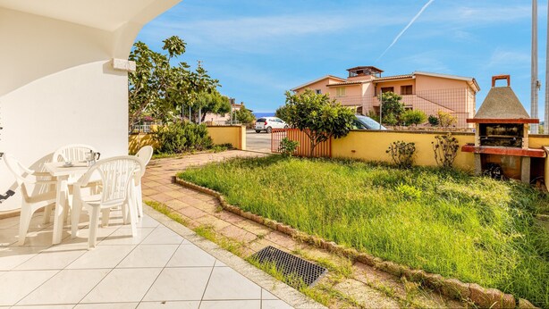A patio with white chairs and a table, a grassy area, and a view of houses and the sea. - Charming Holiday Home Tanaunella appartamento 2 with Ventilators, Balcony, Terraces & Sea View; Parking Available, Pets Allowed (tanaunella)
