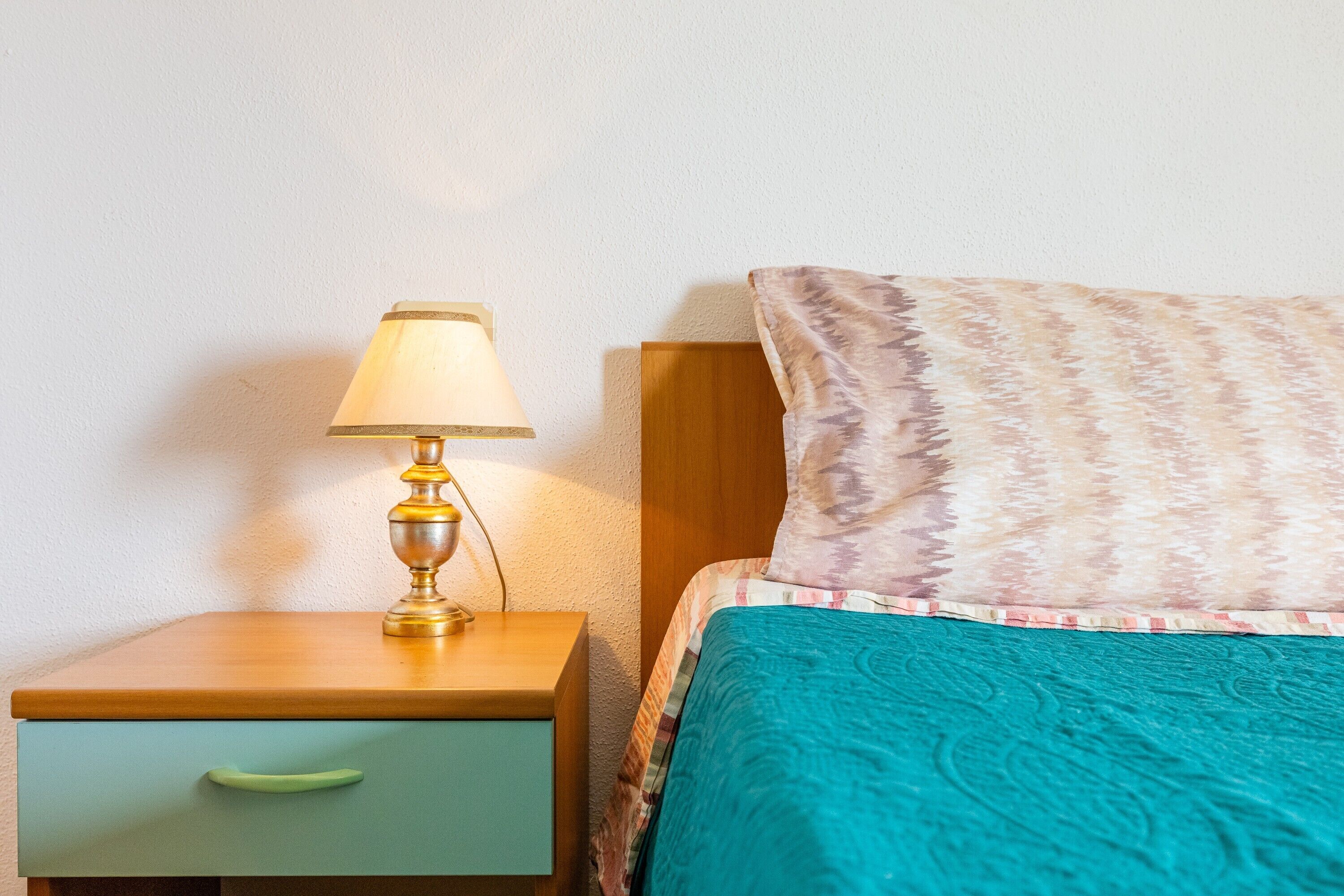 A bed with a patterned pillow, a teal bedspread, and a bedside lamp on a wooden nightstand with a drawer.