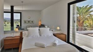 Une chambre moderne avec un grand lit, deux tables de chevet, une commode, une fenêtre donnant sur un paysage verdoyant et un balcon.