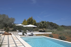 Ein Schwimmbecken mit Liegestühlen und Sonnenschirmen, umgeben von Grünpflanzen und blauem Himmel.
