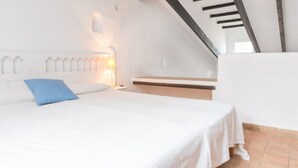 Ein Schlafzimmer mit einem Bett, einem Nachttisch, einem blauen Kopfkissen, einem weißen Kopfteil, einer schrägen Decke und einem Fenster.