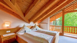 A wooden cabin with a high ceiling, a bed with pillows, a nightstand, and a window with a view of greenery.